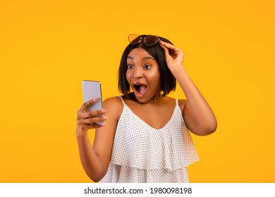 Shocked Black Lady In Summer Outfit Looking At Smartphone In Excitement, Lifting Her Sunglasses On Orange Studio Background. Surprised African American Woman Learning About Online Summertime Sale