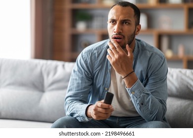 Shocked Black Man With Remote Controller In Hand Sitting On Couch At Home, Confused Young African American Guy Watching Tv Show In Living Room, Emotionally Reacting To Shocking Content, Copy Space
