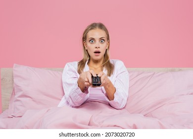 Shocked Blonde Woman In Pajamas Holding Remote Controller On Bed Isolated On Pink