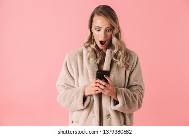 Shocked Blonde Woman Wearing In Fur Coat Using Smartphone With Open Mouth Over Pink Background