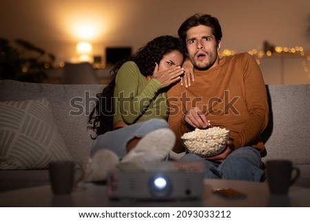 Shocked Boyfriend And Girlfriend Watching Spooky Movie, Lady Covering Eyes In Shock While Sitting Near Projector And Eating Popcorn In Living Room At Home. Front View, Selective Focus