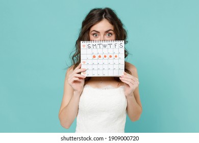 Shocked bride young woman 20s in white wedding dress cover face with female periods calendar for checking menstruation days isolated on blue turquoise background. Ceremony celebration party concept