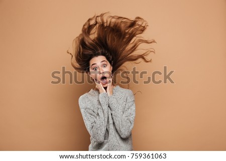 Shocked brunette woman with funny hairstyle touching her face and looking at camera, isolated over beige background