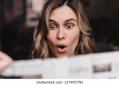 Shocked casual woman reading newspaper and look surprised while standing at home. Shocked curly girl look amazed, when read news in newspaper. Big eyes and open mouth.
