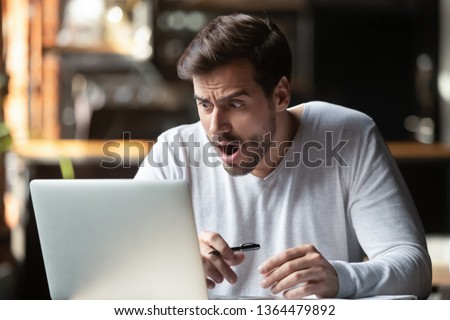 Shocked caucasian man millennial businessman sitting alone at table looking at laptop screen feels surprised and worried opening mouth making big eyes. Unbelievable things awful news problems concept