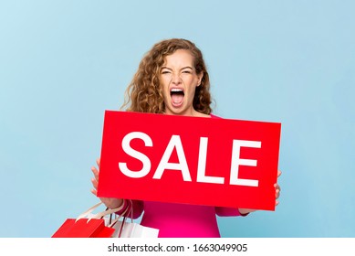 Shocked Caucasian Woman Carrying Shopping Bags Showing Red Sale Sign Isolated On Light Blue Bluebackground