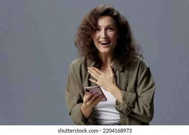 Shocked Cheerful Excited Curly Woman In Casual Khaki Shirt Received Message About Major Sale Of NFT Project Using Phone Posing Isolated On Gray Background. Social Media Influencer Concept. Copy Space