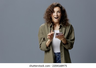 Shocked Cheerful Excited Curly Woman In Casual Khaki Shirt Received Message About Major Sale Of NFT Project Using Phone Posing Isolated On Gray Background. Social Media Influencer Concept. Copy Space