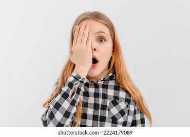 Shocked And Concerned Teen Girl Open Mouth And Covers Half Of Face With Palm, Looks With One Eye At Camera, Stands Over White Studio Background