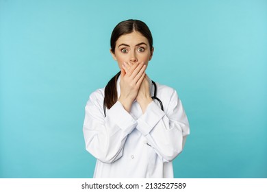 Shocked And Concerned Woman Doctor, Clinic Worker, Covering Mouth With Hands, Looking Startled At Camera, Standing Over Blue Background