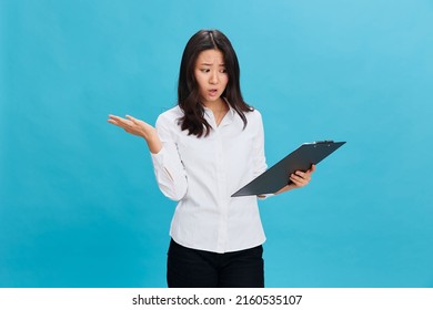 Shocked Confused Cute Asian Businesswoman In Classic Office Dress Code Holds Folder Tablet With Bad Exam Results Posing Isolated On Blue Studio Background. Business Offer. Job Interview Concept