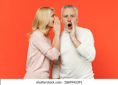 Shocked couple friends elderly gray-haired man blonde woman wearing white pink casual clothes whispering secret behind hand, sharing news isolated on bright orange color background studio portrait