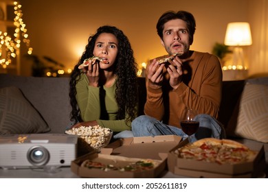 Shocked Couple Watching Horror Movie And Eating Pizza Using Home Cinema Projector Sitting On Sofa In Living Room Indoors At Night. Family Entertainment And Weekend Leisure Concept