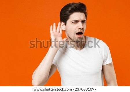 Shocked curious young man 20s in basic casual white blank t-shirt standing try to hear you overhear listening intently looking camera isolated on bright orange colour wall background studio portrait