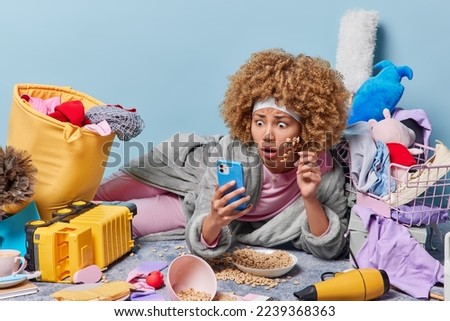 Shocked curly haired impressed woman stares in smartphone screen rects to amazing news dressed in robe feels lazy surrounded by different house poses at messy room isolated over blue background
