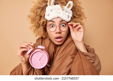 Shocked curly haired woman wakes up after seeing nightmare stares bugged eyes overslept work holds alarmclock keeps hand on rim of spectacles wears sleepmask on forehead wrapped in soft blanket