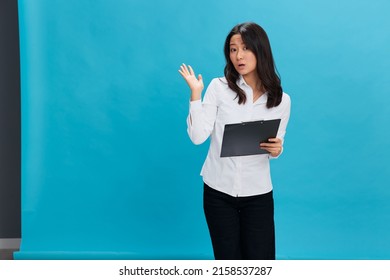 Shocked Cute Asian Businesswoman In Classic Office Dress Code Holds Folder Tablet Tells Secret Posing Isolated On Over Blue Studio Background. Cool Business Offer. Job Interview Concept