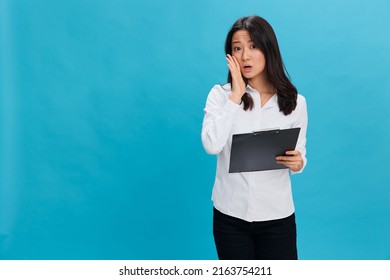 Shocked Cute Asian Businesswoman In Classic Office Dress Code Holds Folder Tablet Tells Secret Posing Isolated On Over Blue Studio Background. Cool Business Offer. Job Interview Concept