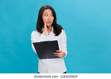 Shocked Cute Asian Businesswoman In Classic Office Dress Code Holds Folder Tablet Tells Secret Posing Isolated On Over Blue Studio Background. Cool Business Offer. Job Interview Concept