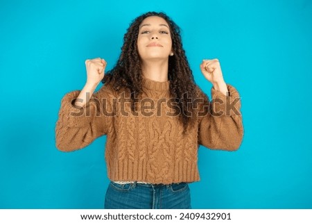 Shocked ecstatic Beautiful arab girl with curly hair wearing knitted sweater win luck lottery raise hands up shout yea