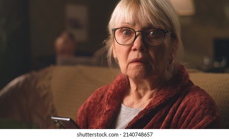 Shocked elderly female in glasses looking around after reading terrifying news on mobile phone while sitting on sofa in living room at home