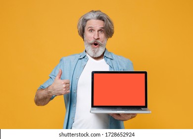 Shocked Elderly Gray-haired Mustache Bearded Man In Casual Blue Shirt Posing Isolated On Yellow Background. People Lifestyle Concept. Hold Laptop Computer With Blank Empty Screen, Showing Thumb Up