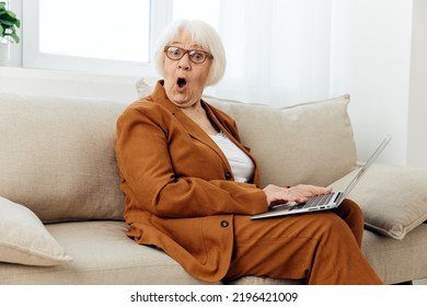 A Shocked Elderly Woman Is Sitting With Her Mouth Wide Open From Severe Shock Holding A Laptop On Her Lap Sitting On A Comfortable Sofa