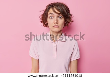 Shocked embarrassed woman with short hairstyle stares bugged eyes feels anxious drressed in casual t shirt isolated over pink background looks in disbelief hears something impressive. Human reactions