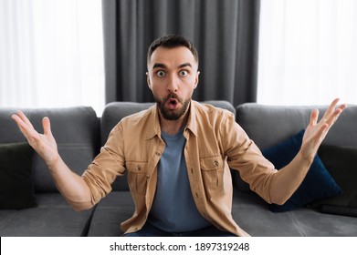 Shocked Emotional Man. Surprised Caucasian Guy Sits On The Couch, Looking Directly At The Camera, Got An Unexpected News, Gestures With His Hands