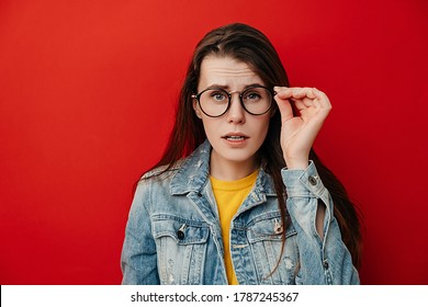 Shocked emotional young woman keeps hand on rim of spectacles, dressed in denim jacket, gazes with stupefaction, being amazed by terrifying news, isolated on red studio background. Reaction concept