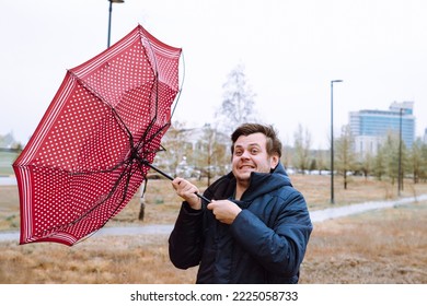 Shocked European White Young Male Man Fool Around With Umbrella In City Park, Strong Storm Wind, Rain And Bad Weather. Wind Broke Umbrella. Autumn Waist Up Lifestyle Portrait.