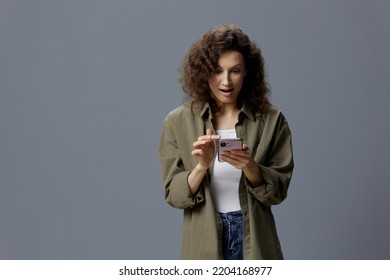 Shocked Excited Confused Curly Beautiful Woman In Casual Khaki Green Shirt Get Shock Info From Message Using Phone Posing Isolated On Gray Blue Background. Social Media Influencer Concept. Copy Space