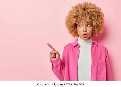 Shocked Excited Woman With Curly Hair Points Finger On Left Shows Blank Space For Your Promo Opens Mouth From Amazement Wears Turtleneck And Shirt Isolated Over Pink Background. Omg Look There