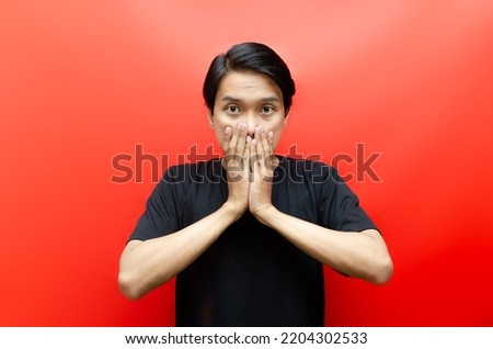 Shocked face of Asian man in black shirt on red background. man feeling embarassed shocked with hand covering mouth.