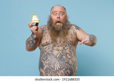 Shocked fat pudge obese chubby overweight man has tattooed big belly hold sweet pastries cake pointing index finger on stomach isolated on blue background. Weight loss obesity unhealthy diet concept