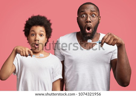 Shocked father and son hold toothbrushes, brush teeth and hurry as late for work or school, overslept, isolated over pink background. Dark skinned African male adult and teenager in bathroom