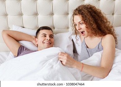 Shocked female looks under white blanket, being surprised with size of boyfriend`s genitails or sees man`s erection. Satisfied couple have good relationships. Intimate and lifestyle concept.