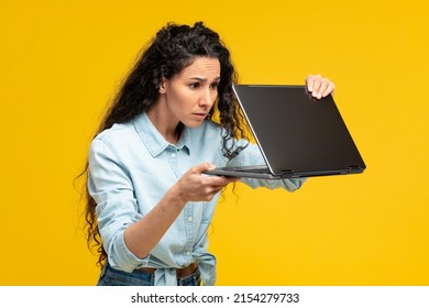 Shocked Frightened Woman With Curly Hair Holding Pc Looking Furtively At Half Closed Laptop, Upset Scared Of Error Or Software Failure, Blue Screen Of Death. Lady Using Computer On Yellow Orange Wall