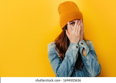 Shocked frightened young woman peeks through fingers, stares with widely opened eyes, wears denim jacket and hat, curious female student peek cover hide face in hands, isolated over yellow background - Powered by Shutterstock