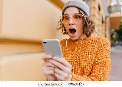 Shocked Girl With Brown Eyes Looking At Phone Screen With Mouth Open. Outdoor Portrait Of Surprised Young Woman With Curly Hair Wearing Yellow Knitted Attire And Holding Smartphone.