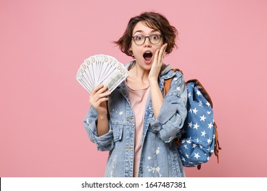 Shocked girl student in denim clothes glasses backpack isolated on pastel pink background. Education in school university college concept. Hold fan of cash money in dollar banknotes put hand on cheek