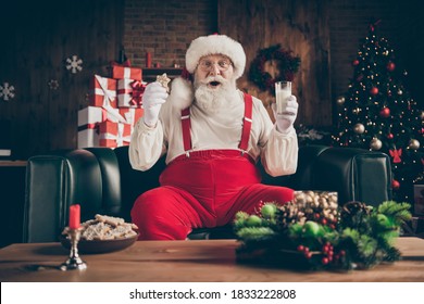 Shocked grey beard santa claus sit cozy couch watch x-mas christmas cinema film impressed drink milk glass eat gingerbread cookie wear red cap headwear in house indoors noel ornament