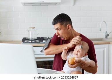 Shocked Handsome Man Wearing Maroon Casual T Shirt Drops Smart Phone While Sitting At Table In Kitchen And Feeding Little Daughter Or Son, Scared Father With Baby Boy Or Girl.