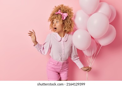 Shocked Impressive Curly Woman Dressed In Fashionable Outfit Notices Something Breathtaking Away Holds Bunch Of Inflated Helium Balloons Has Univeristy Leaving Party Isolated Over Pink Background.