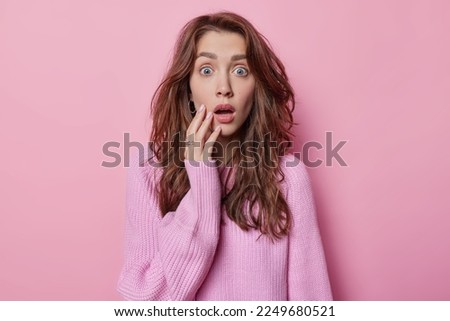 Shocked impressive woman feels stunned keeps mouth opened holds breath from amazement has long wavy hair dressed in knitted jumper reacts to something impressive isolated on pink background.