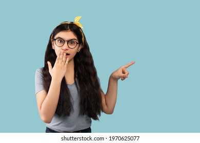 Shocked Indian Adolescent Standing With Open Mouth, Pointing At Empty Space In Disbelief, Blue Studio Background. Overwhelmed Teen Girl Offering Enormous Sale Or Promotion