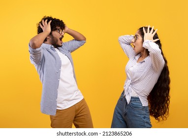 Shocked indian lady and her boyfriend looking at each other with open mouths, feeling surprised and touching heads on yellow studio background. Young couple screaming WOW or OMG