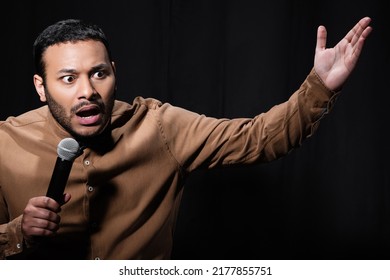 Shocked Indian Stand Up Comedian Telling Joke Into Microphone On Dark Stage Isolated On Black