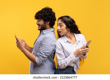 Shocked indian woman spying on her smiling boyfriend who using smartphone, chatting or scrolling social media news feed. Young couple standing back to back over yellow studio background