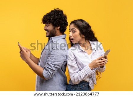 Shocked indian woman spying on her smiling boyfriend who using smartphone, chatting or scrolling social media news feed. Young couple standing back to back over yellow studio background